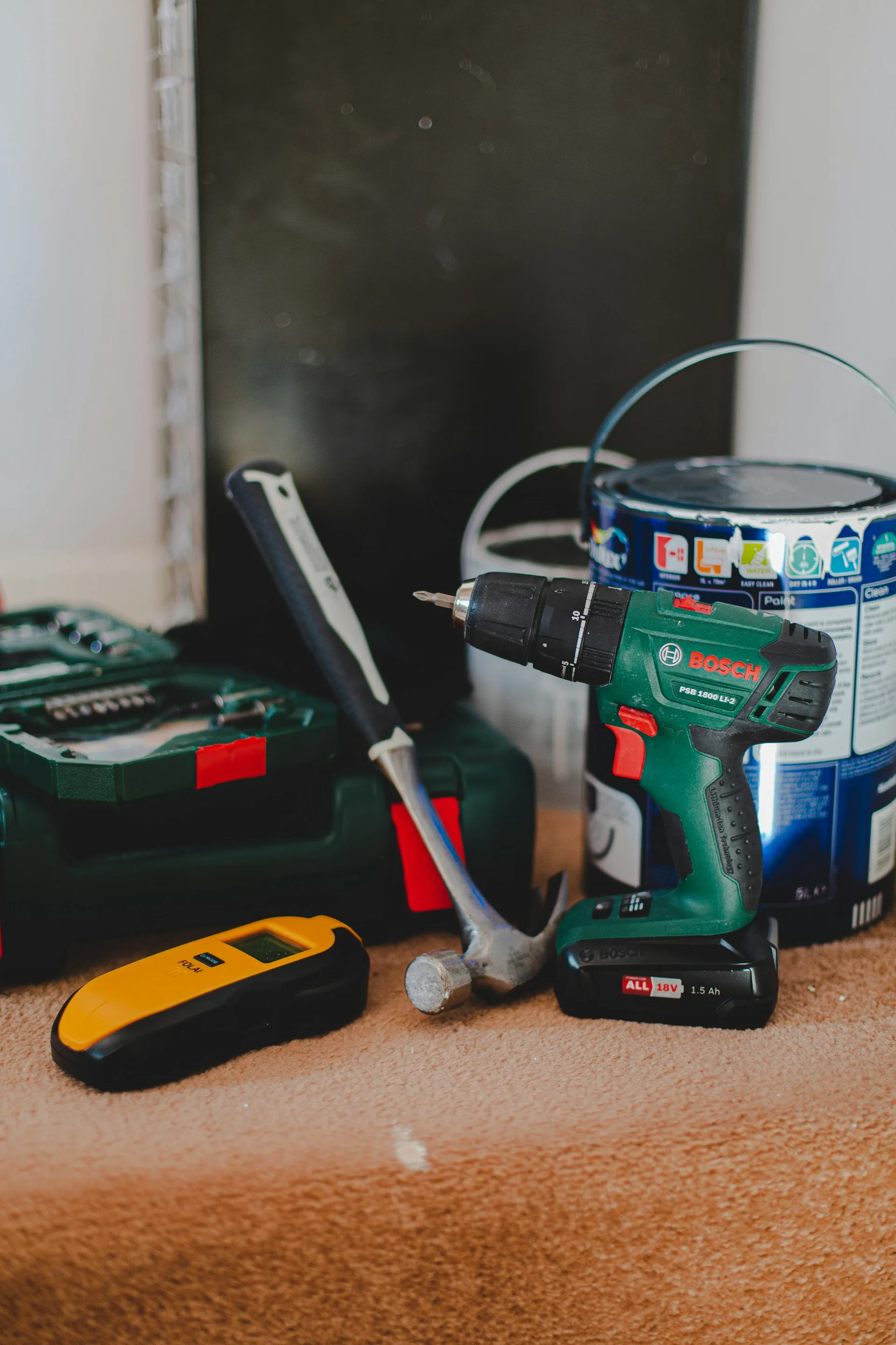 Cordless drill, claw hammer, stud detector and a tin of paint on a dust sheet